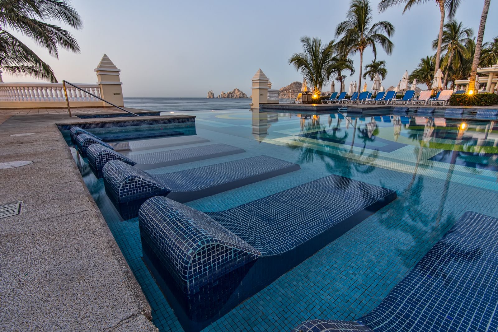Resort pool with in-water loungers and palm trees in Cabo San Lucas, with the Land's End rocks on the horizon