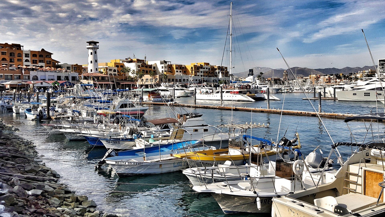 Fishing boats and yachts docked at the Cabo San Lucas marina with the lighthouse and waterfront buildings behind