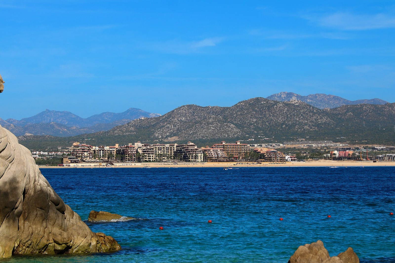 Calm blue water of Médano Beach with hotels along the sand and mountains behind, seen from the rocks