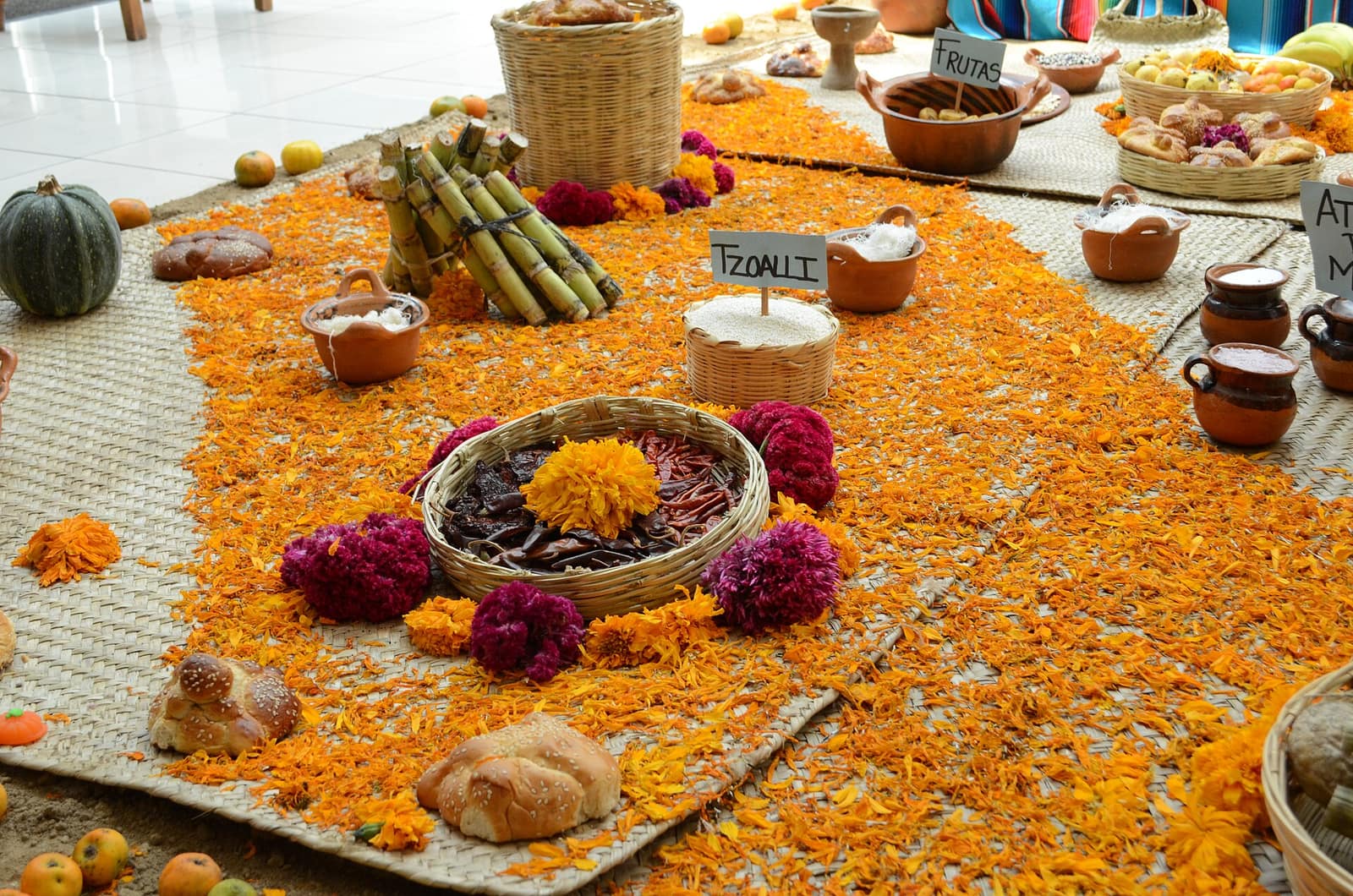 A floor covered in orange marigold petals with bread, fruit, and clay pots arranged as Día de los Muertos offerings