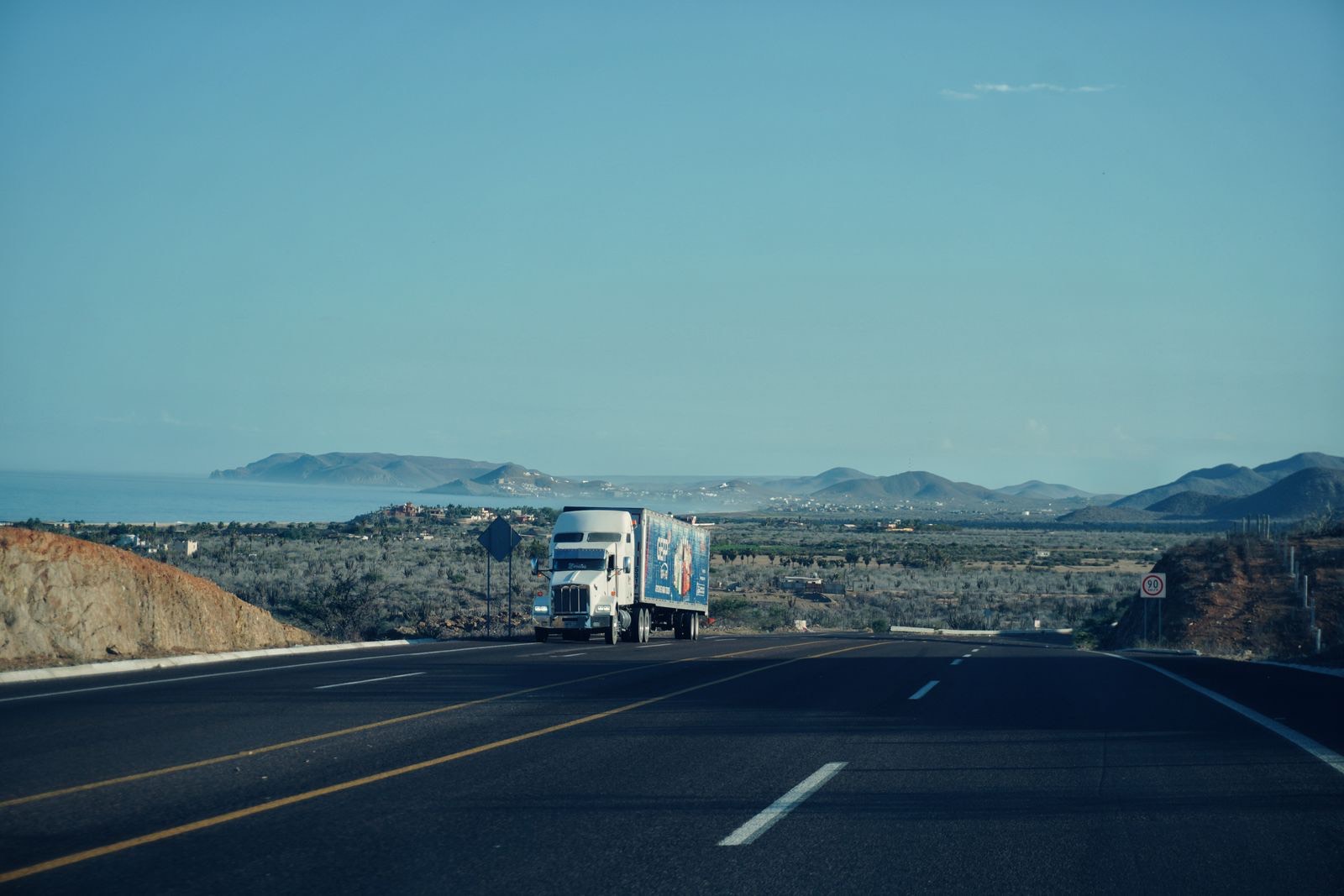 Semi truck on a two-lane desert highway with mountains and the sea in the distance