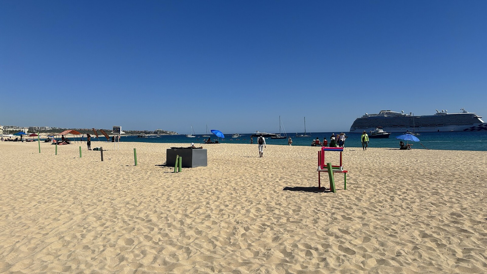 Wide sandy Médano Beach in Cabo San Lucas with calm bay water, anchored boats, and a cruise ship offshore