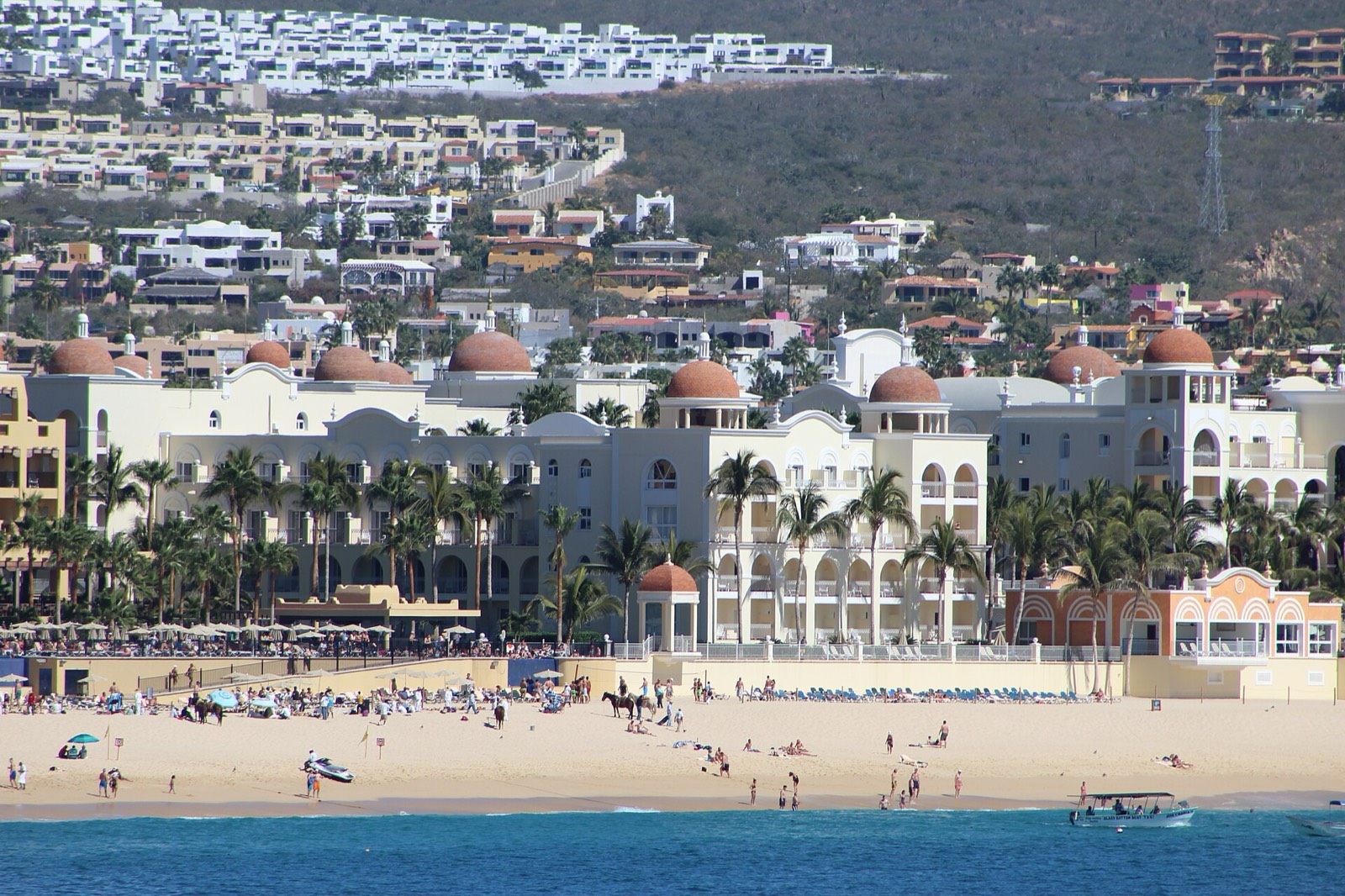 Crowded sandy beach and calm water in front of a large white resort with terracotta domes in Cabo San Lucas